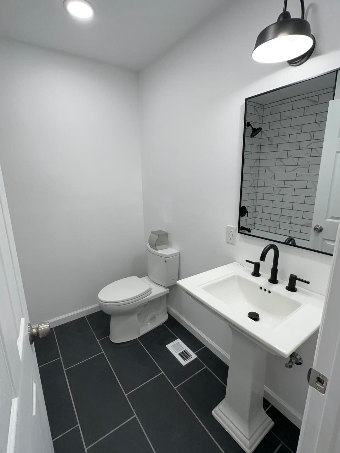 Small modern bathroom with white walls, dark tile floor, toilet, pedestal sink, and mirror light