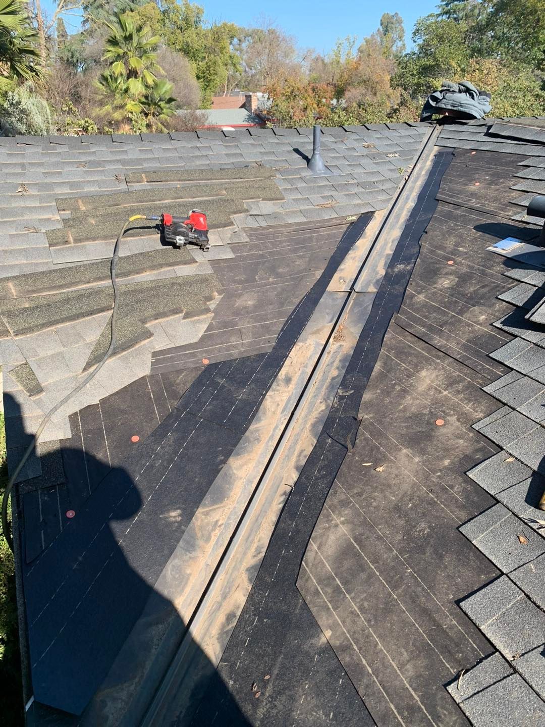 Damaged shingled roof with a long black tarp-covered seam and a small red power tool on the left