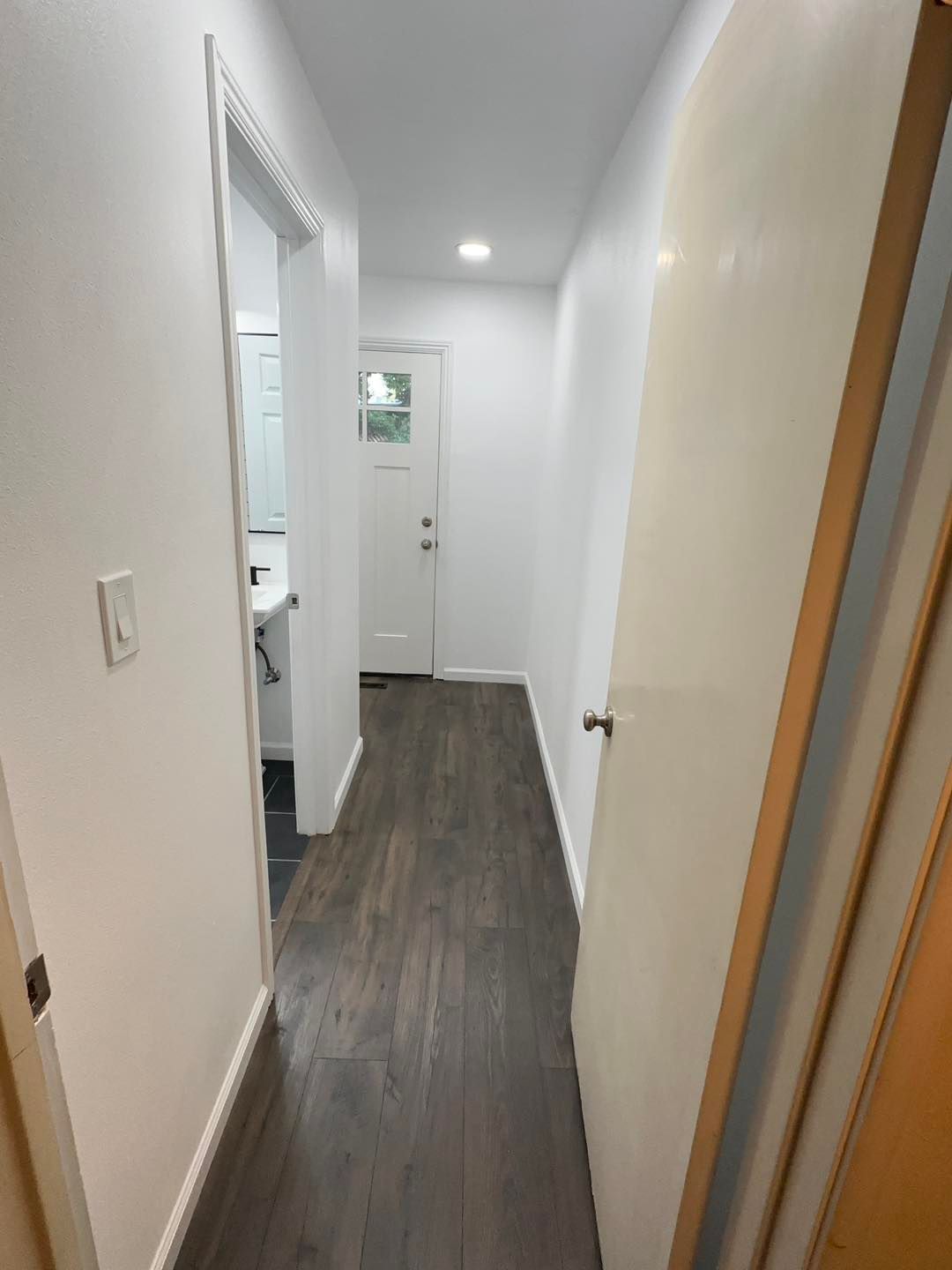Narrow white hallway with dark wood floor, doorway on left, and a door at the far end.