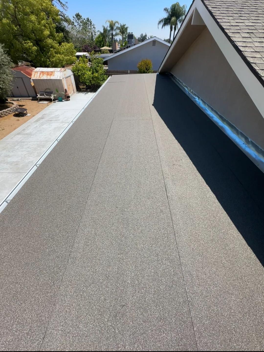 Rooftop under construction with gray roofing material and blue edge trim, viewed beside neighboring houses