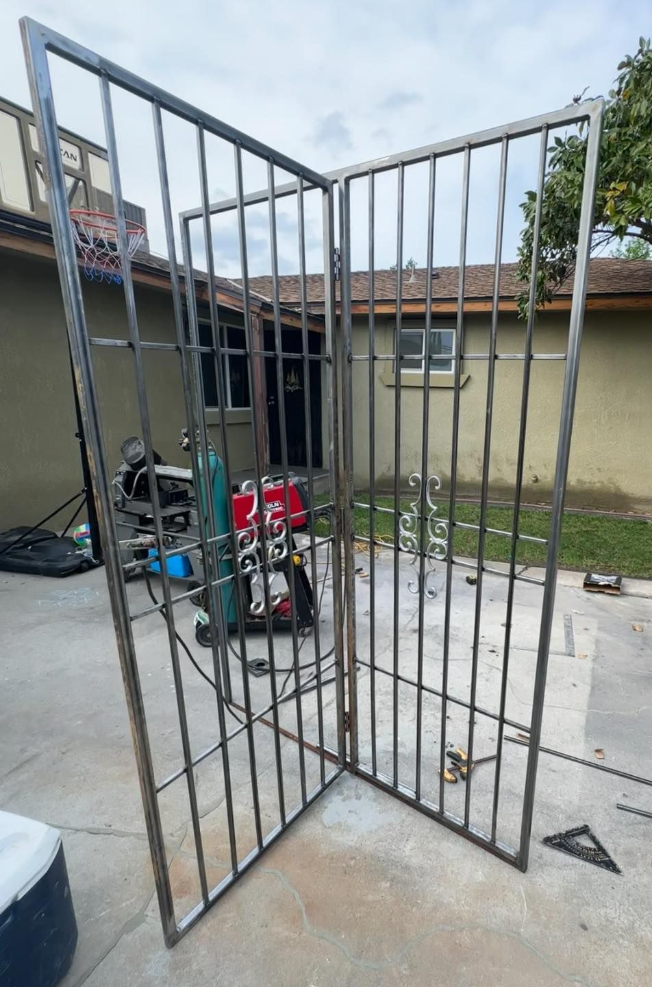 Open metal gate in a concrete courtyard beside a beige building and scattered equipment.