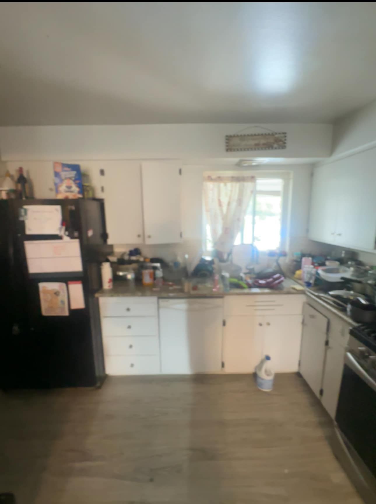 Small kitchen with white cabinets, black fridge, cluttered countertops, and a window over the sink.