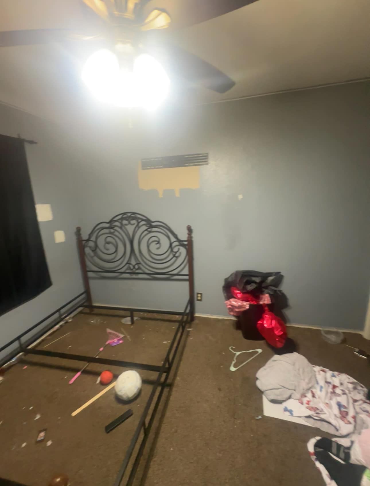 Messy bedroom with a bed frame, balloons, clothes on the floor, and a bright ceiling fan light.