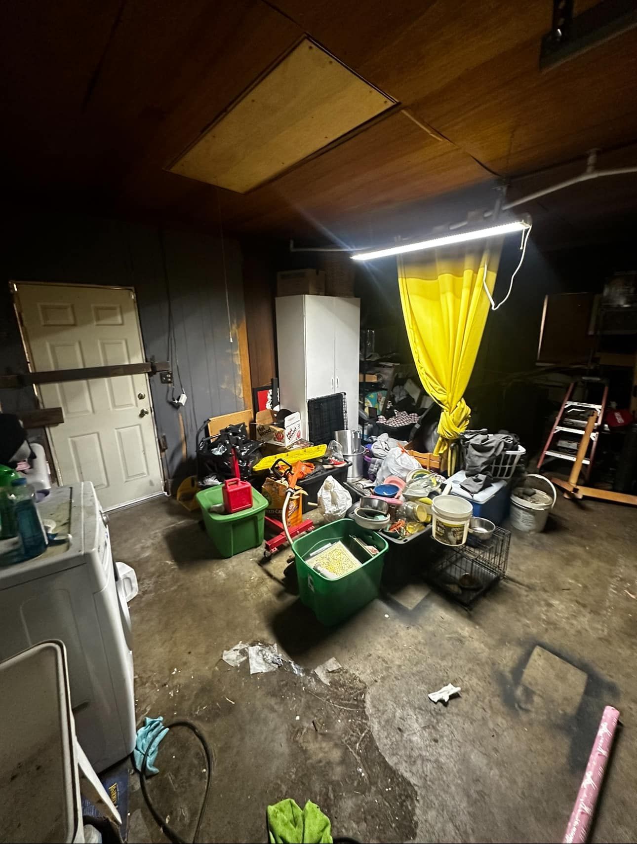 Cluttered garage with piled household items, a yellow curtain, and debris on a wet concrete floor.
