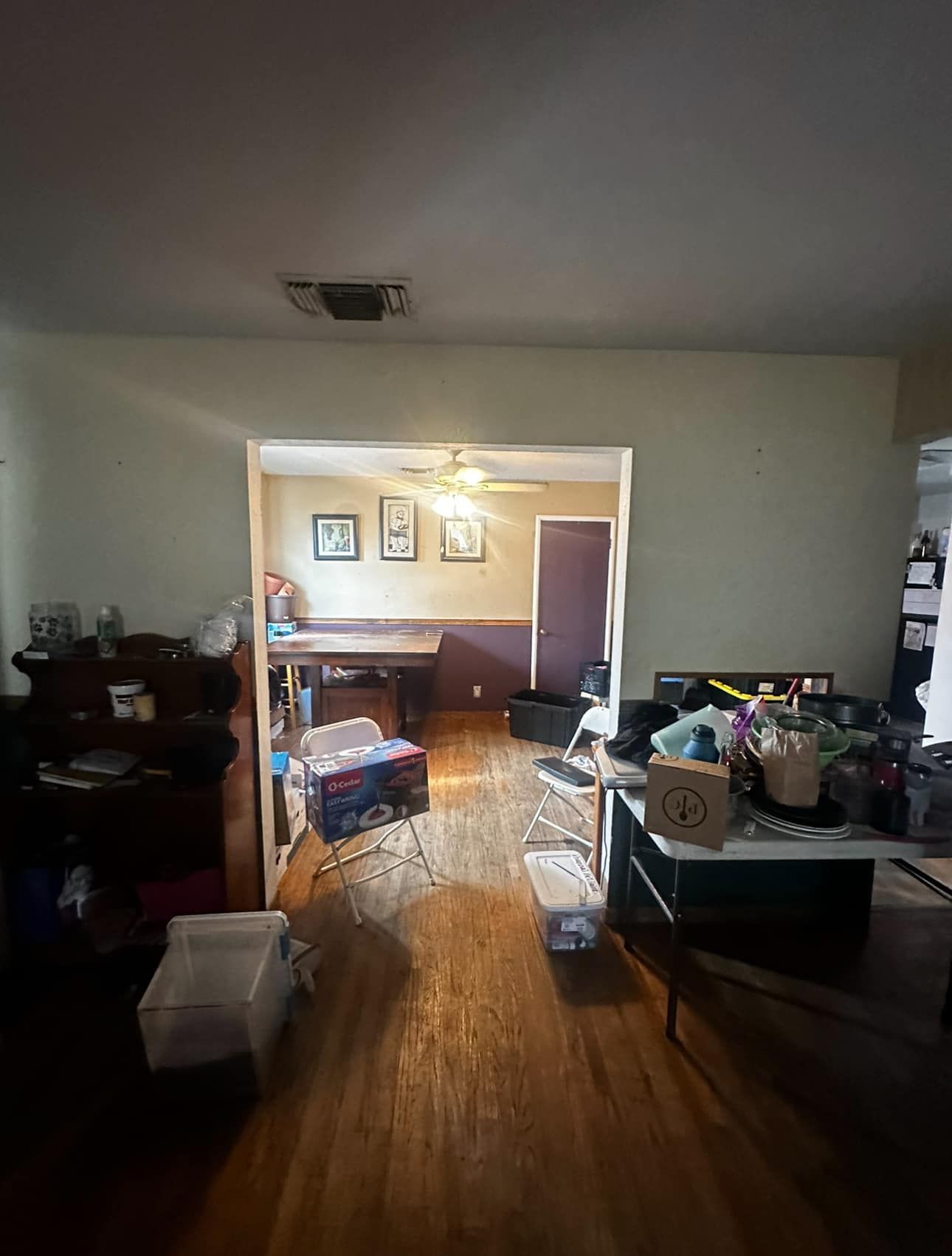 Cluttered living room with wood floors, table, boxes, and a doorway to a lit room beyond.