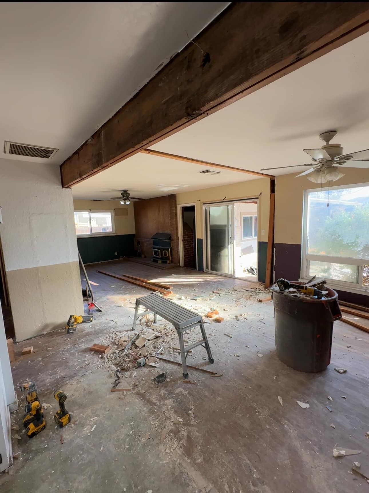 Messy gutted living room with debris, exposed ceiling beam, and large windows overlooking outdoors