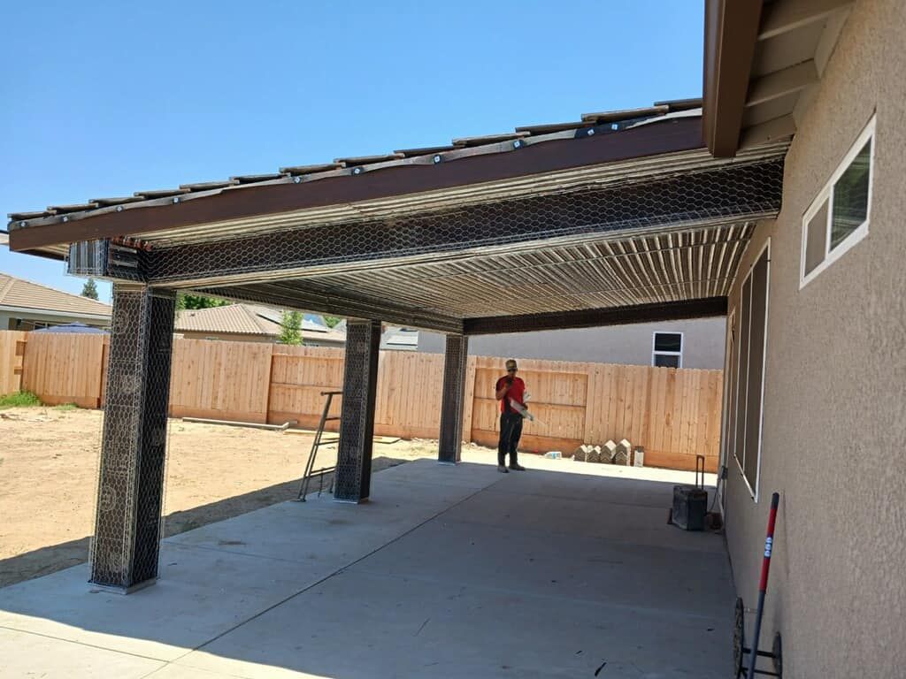Backyard patio with a slatted pergola and a person standing near the fence under a covered concrete slab