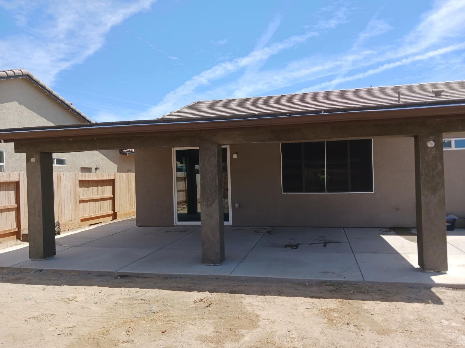 Covered backyard patio with concrete slab, wooden posts, and a windowed wall under a blue sky