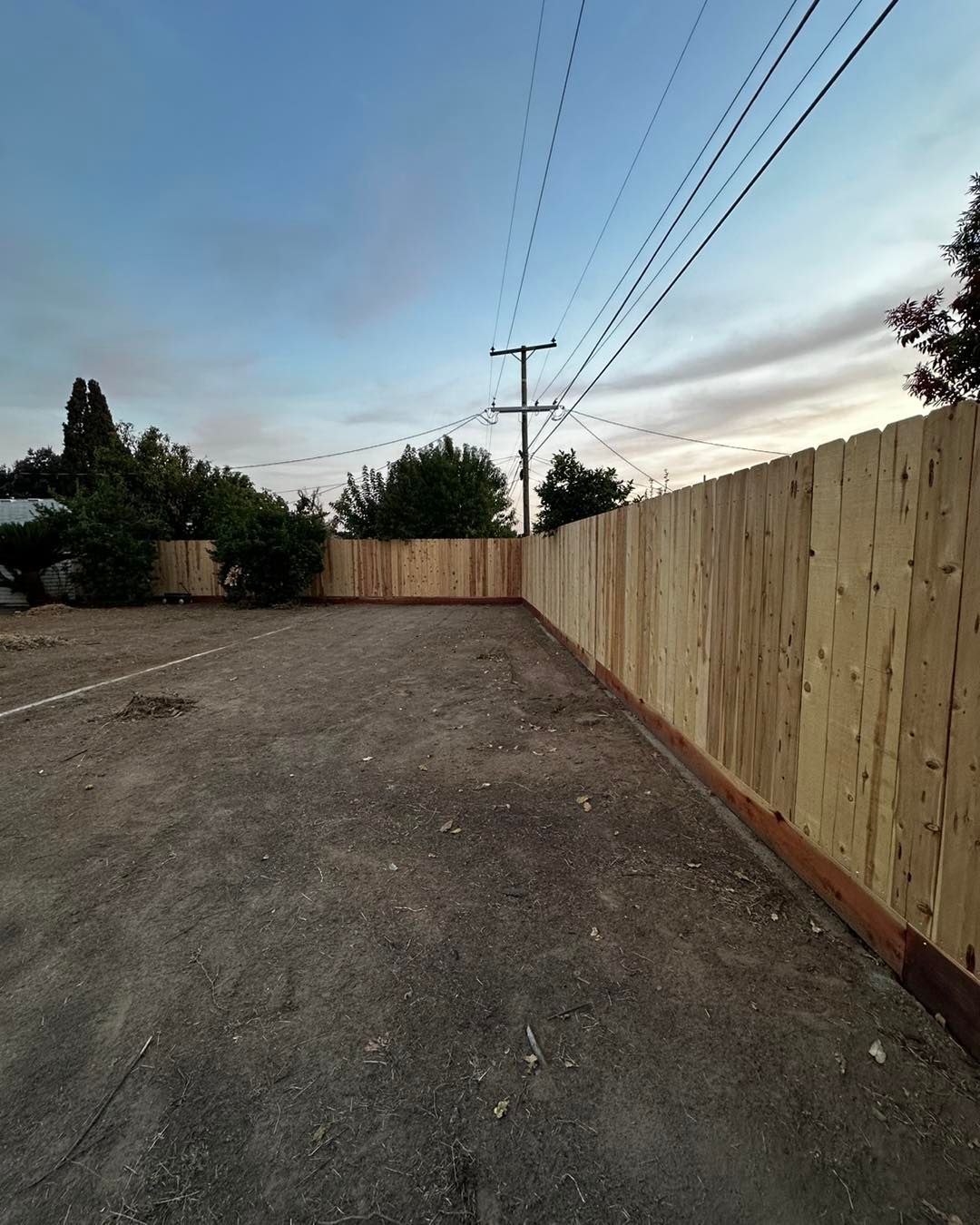 Gravel driveway beside a tall wooden fence under a blue evening sky with power lines overhead