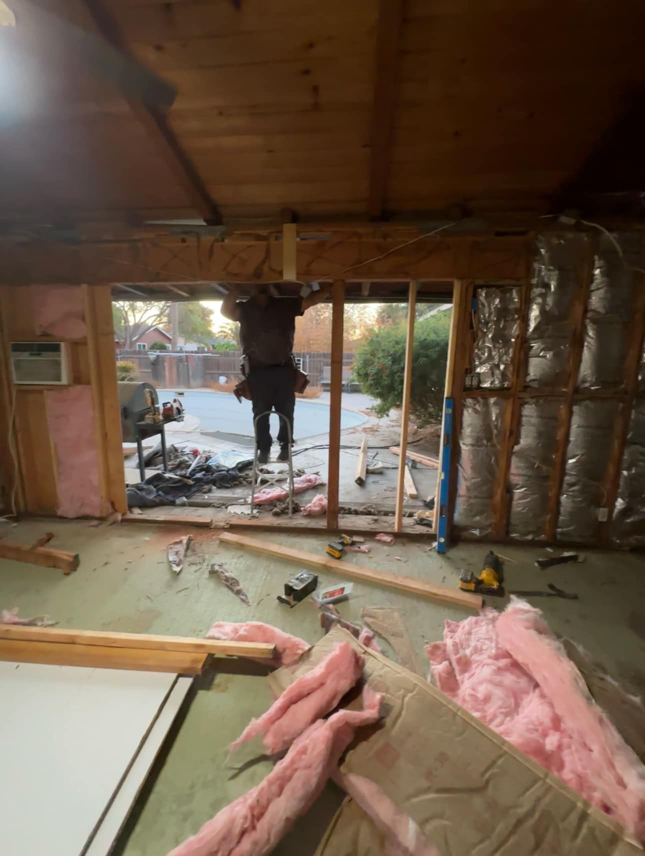 Interior under renovation with exposed walls, pink insulation, and a worker cutting through a large opening to outdoors