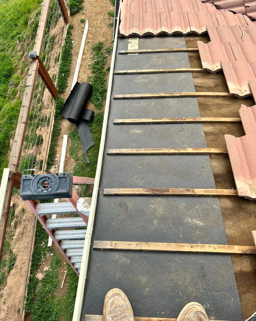 Rooftop under repair with black roofing underlayment, wooden battens, and stacked red tiles along the edge