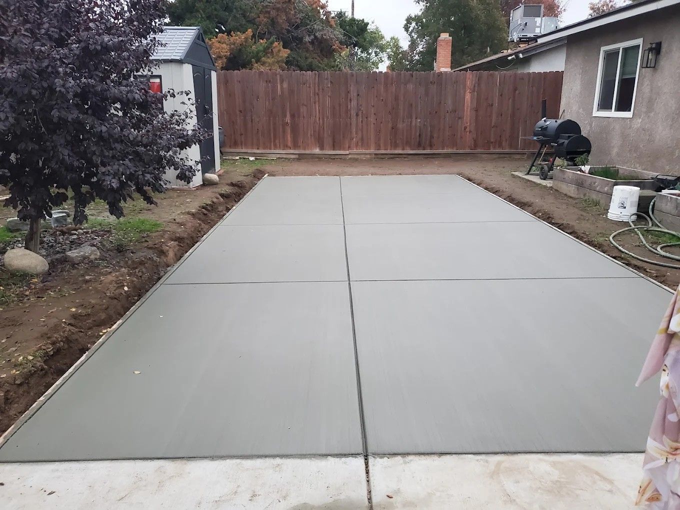 Fresh concrete driveway in a backyard, bordered by dirt and a wooden fence, with a person at the right edge.