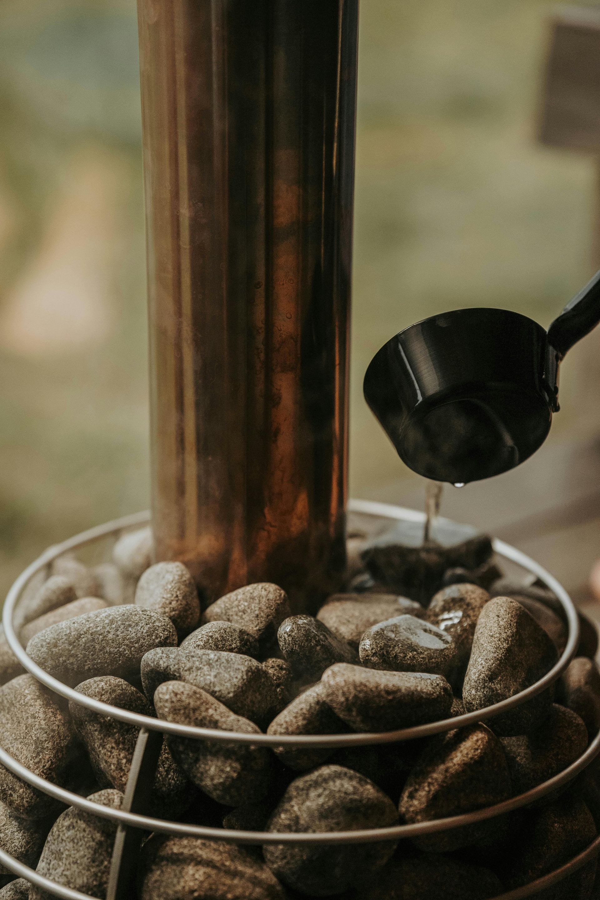 Water being poured from a black ladle onto hot stones surrounding a copper sauna heater.