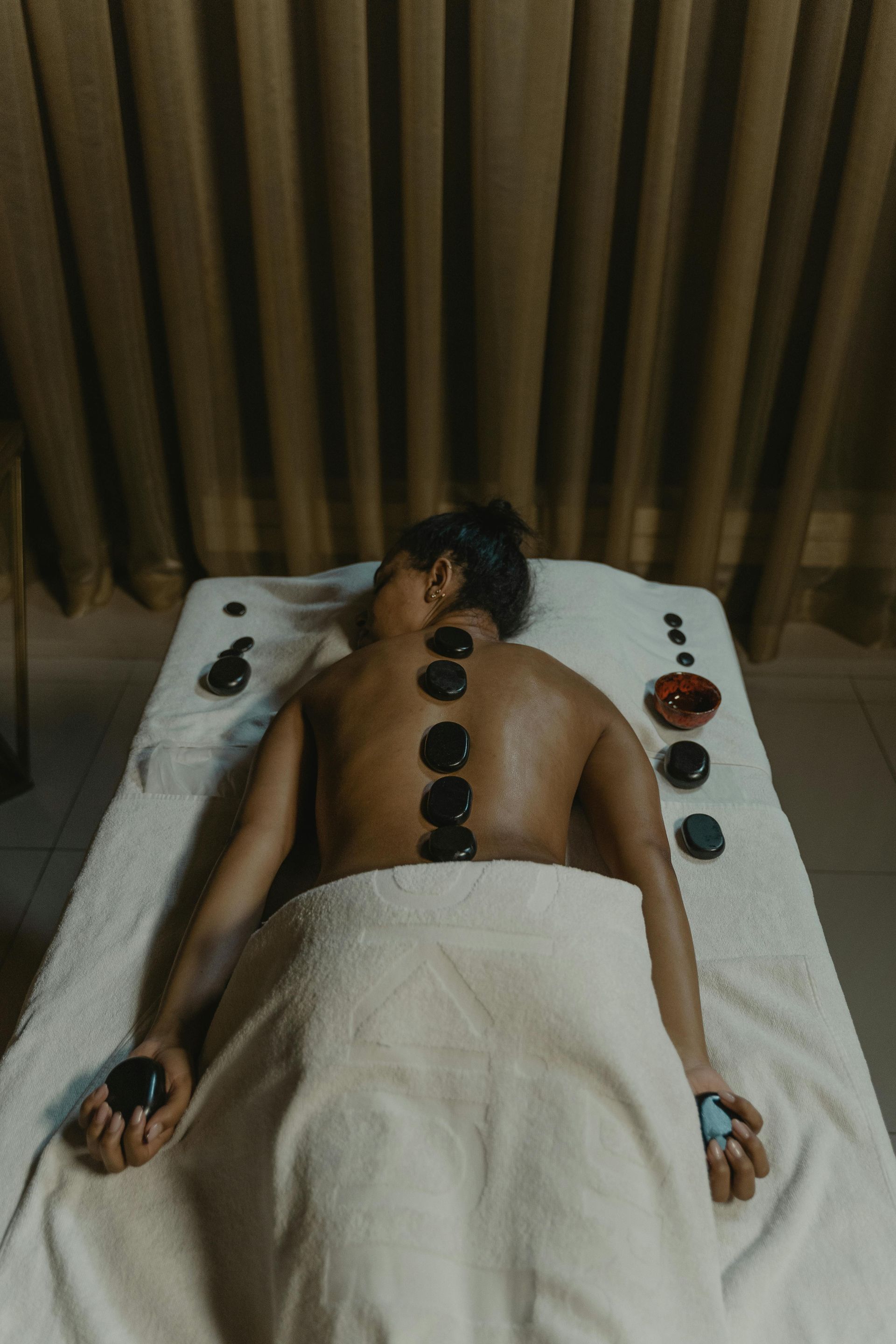 Person receiving a hot stone massage on their back in a spa setting with beige curtains.