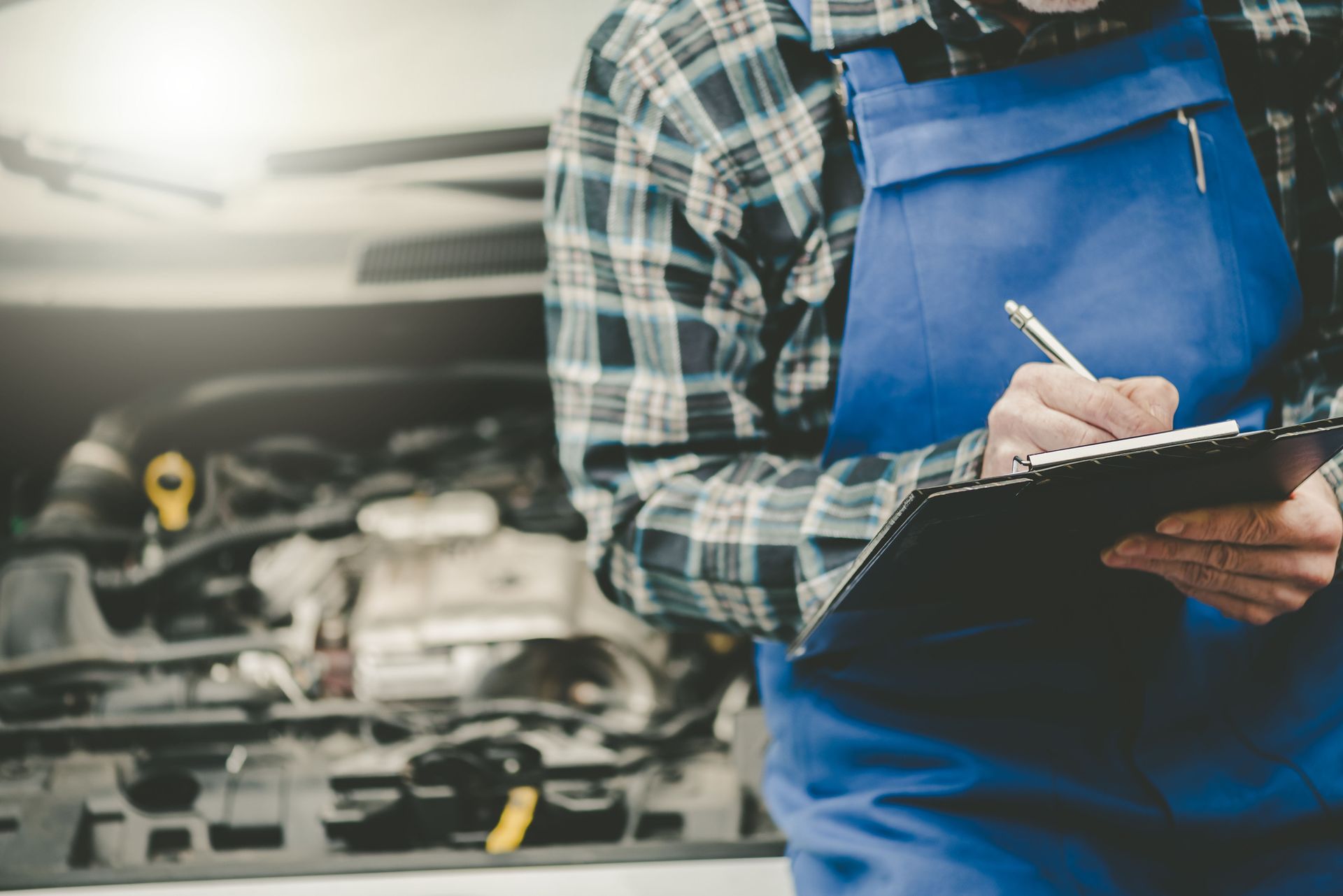 Meccanico in tuta blu e camicia a quadri che scrive su un blocco per appunti, sullo sfondo il motore di un'auto.