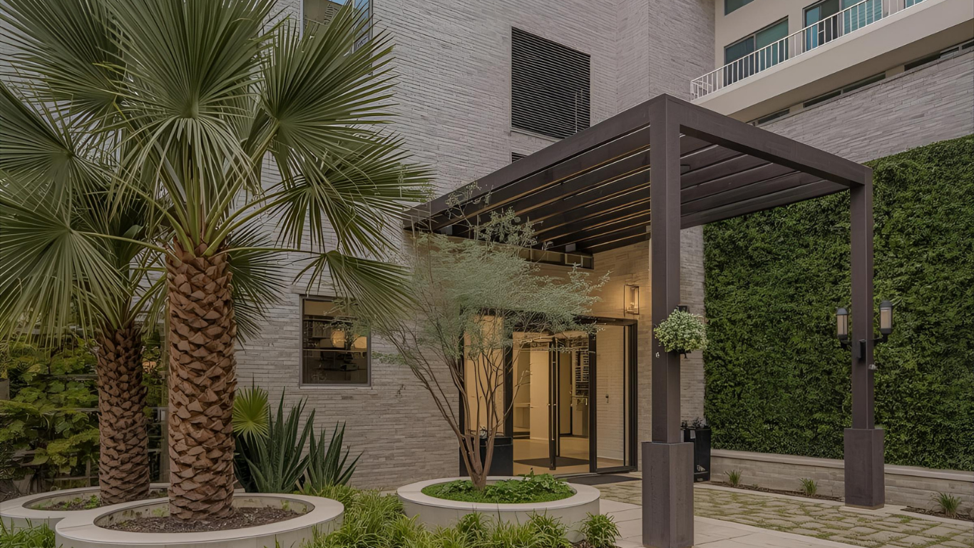 Entrance to a modern building with a pergola, palm trees, and greenery.
