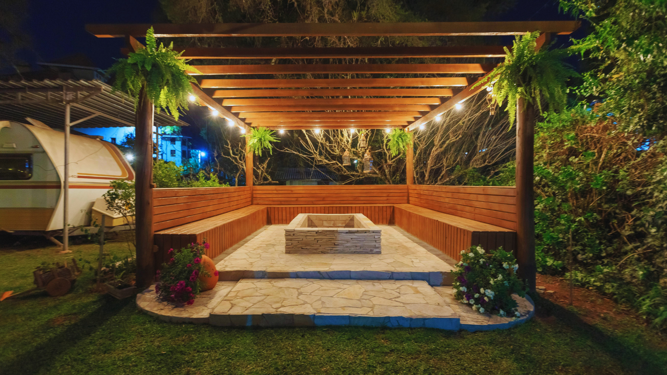 Wooden pergola with benches surrounding a fire pit, lit by string lights, in a backyard.