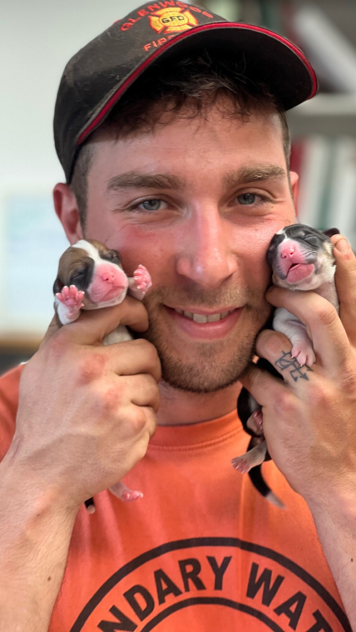 A man in an orange shirt is holding two puppies in his hands.