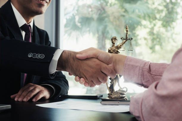 Two people shake hands over desk with documents and Lady Justice statue.