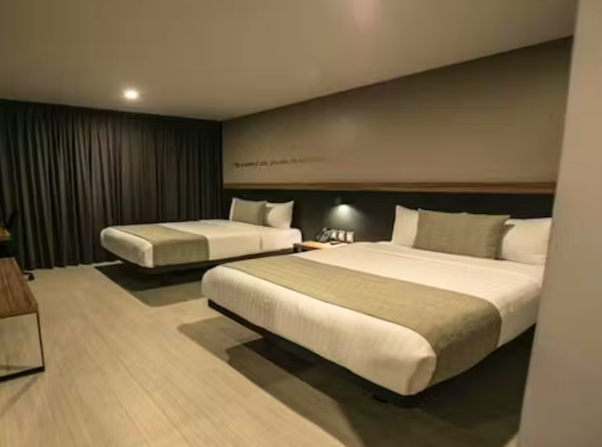 Habitación de hotel con dos camas queen, colores neutros, cortinas oscuras e iluminación cenital.