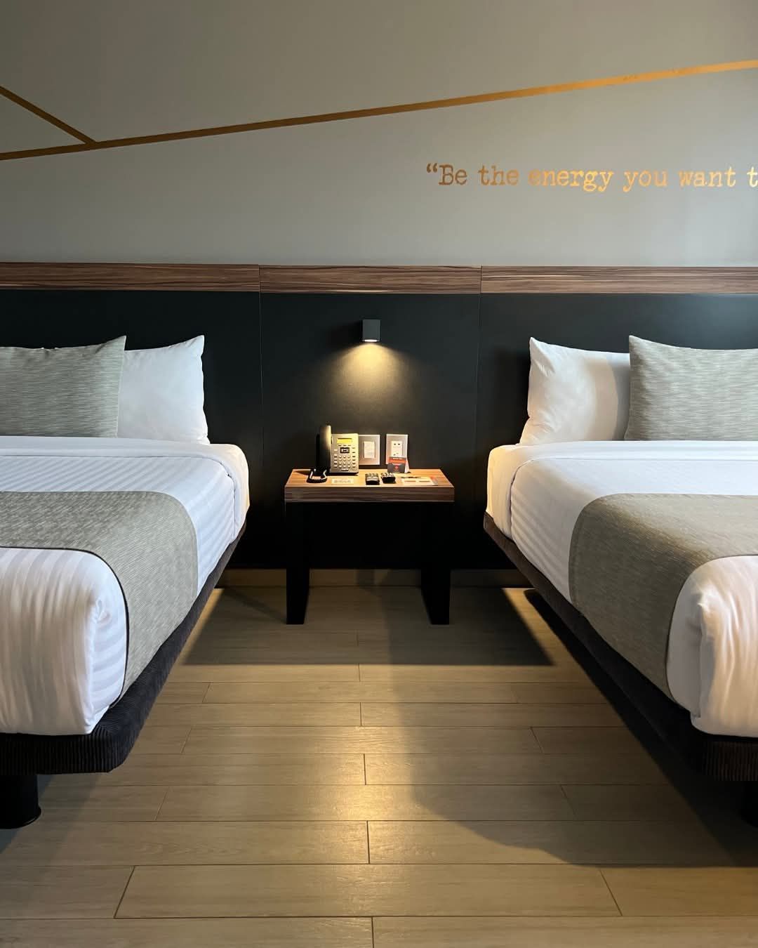 Dos camas en una habitación de hotel, cabeceros oscuros, mesita de noche pequeña, tonos neutros, cita inspiradora en la pared.