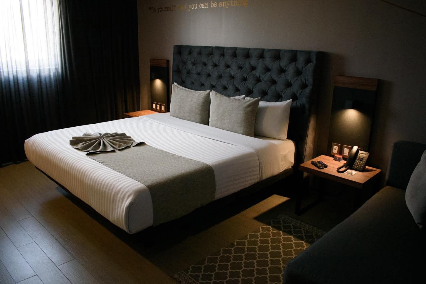 Habitación de hotel con una cama grande, cabecero capitoné y dos mesitas de noche. Tonos oscuros e iluminación concentrada.