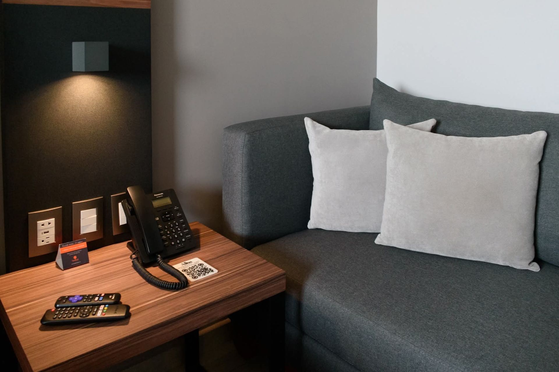 Un pequeño rincón de habitación de hotel con un sofá gris, dos almohadas blancas, una mesa auxiliar de madera con un teléfono, enchufes y una lámpara.