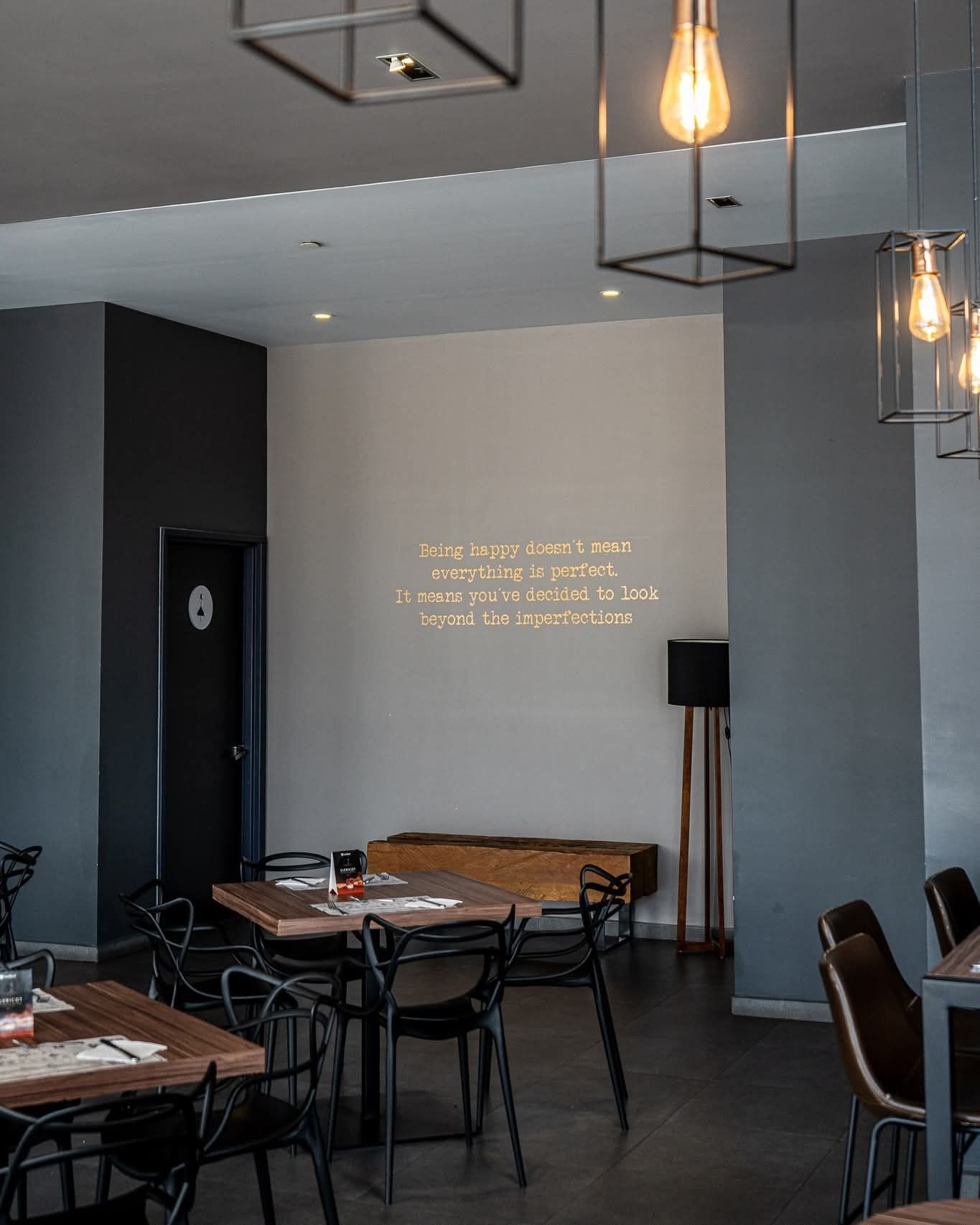 Interior de restaurante con mesas, sillas y luces colgantes; paredes de color gris oscuro y neutro; una cita en la pared.