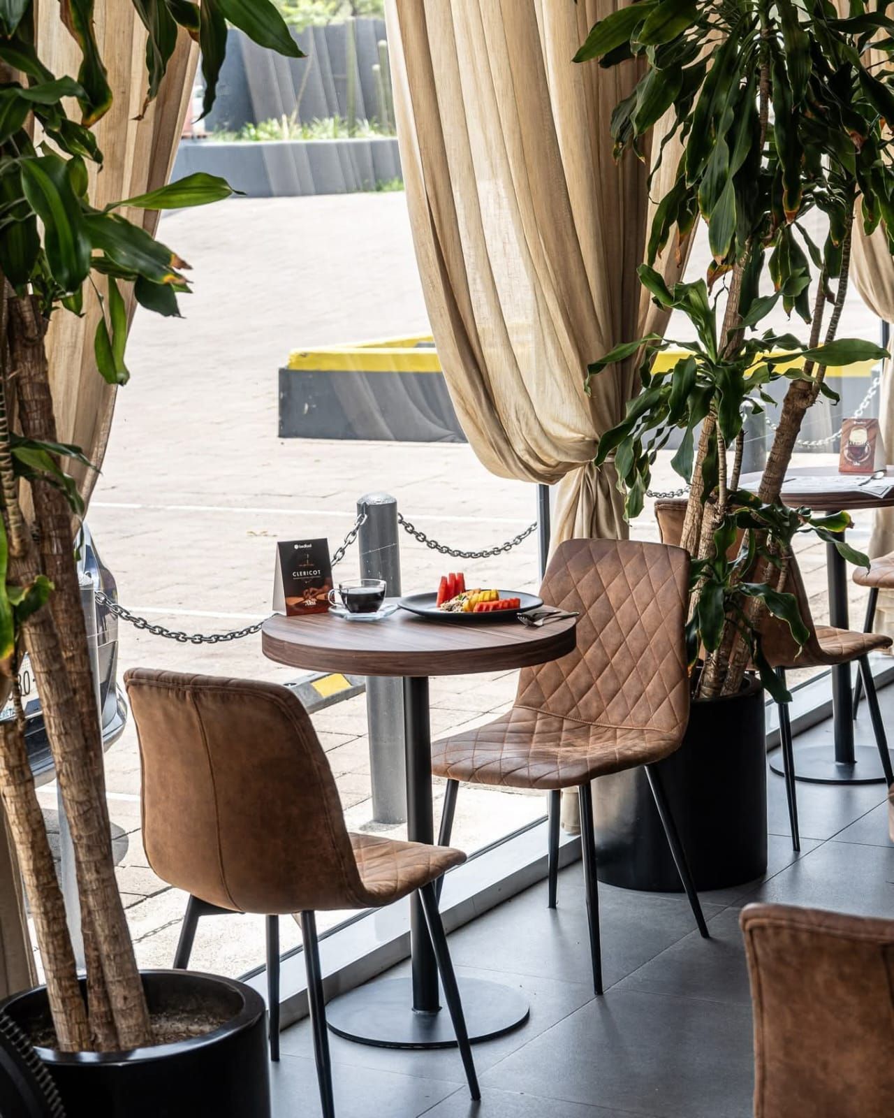 Restaurante con asientos junto a una ventana, con sillas marrones, mesa redonda y plantas.