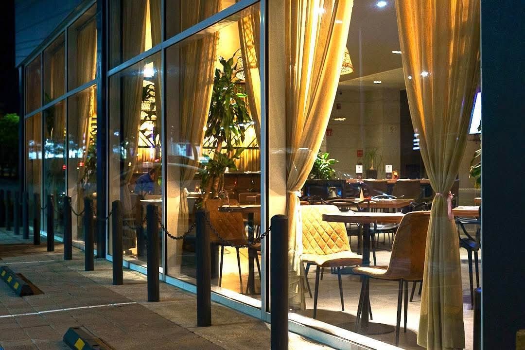 Vista exterior de un restaurante con grandes ventanales de cristal, cortinas doradas y mesas con sillas visibles en el interior.