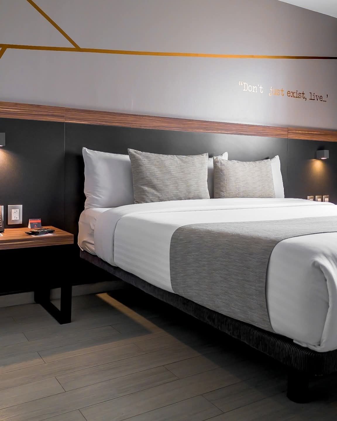 Dormitorio con cama king, cabecera negra, mesita de noche de madera y ropa de cama gris y blanca.