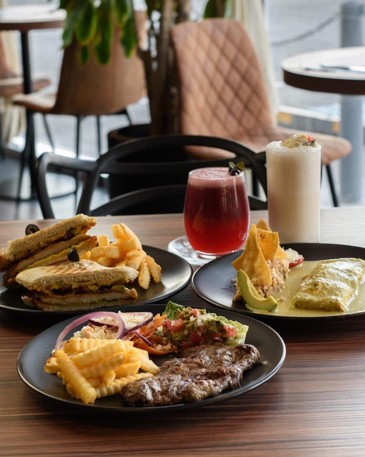 Varios platos de comida y bebidas en una mesa de un café, incluido un filete, un sándwich y cócteles.