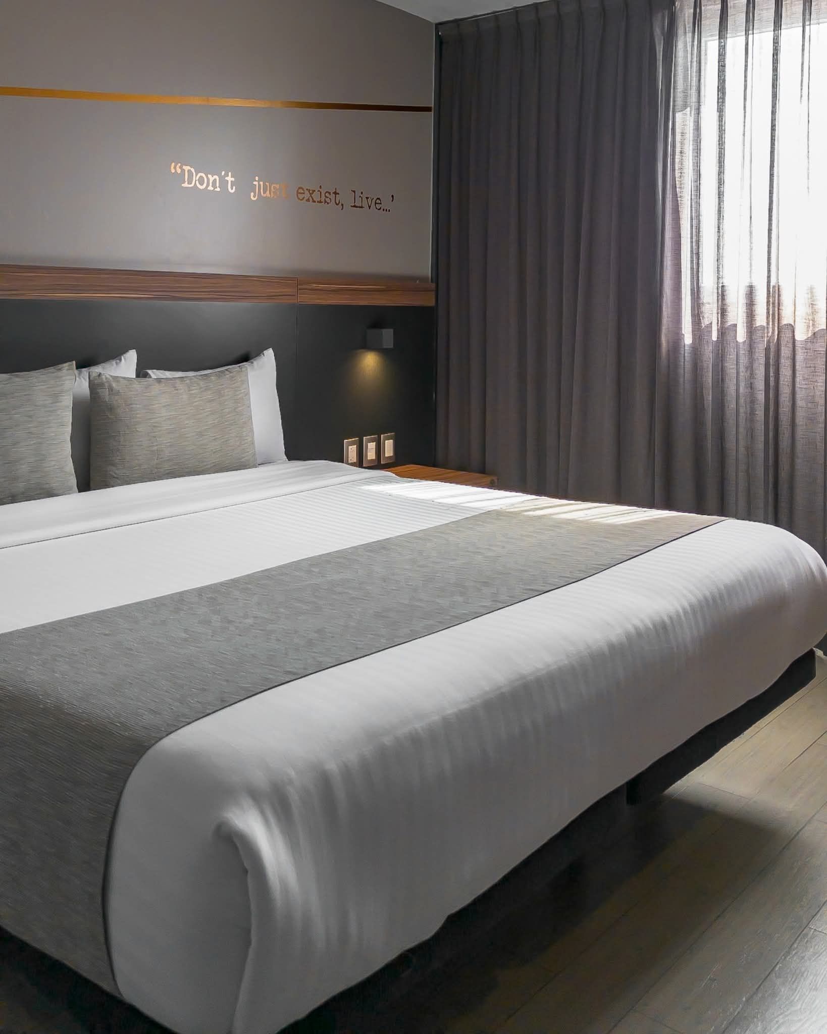 Dormitorio con cama grande, ropa de cama gris y blanca, y una ventana con cortinas grises.