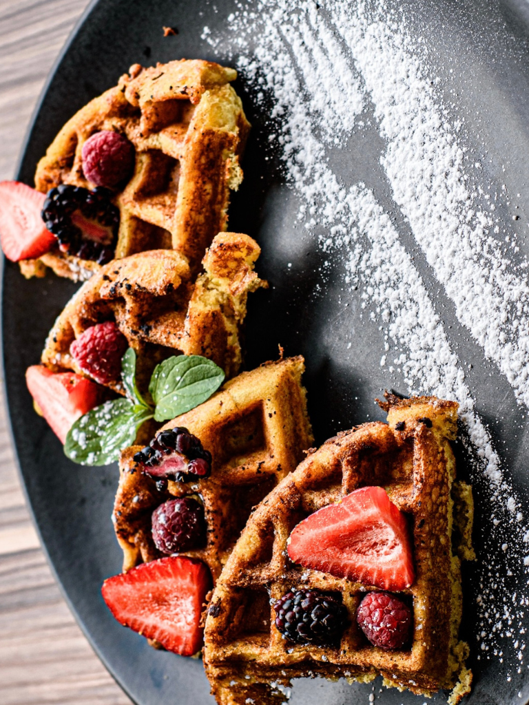 Waffles cubiertos con fresas, arándanos, moras y azúcar en polvo en un plato negro.