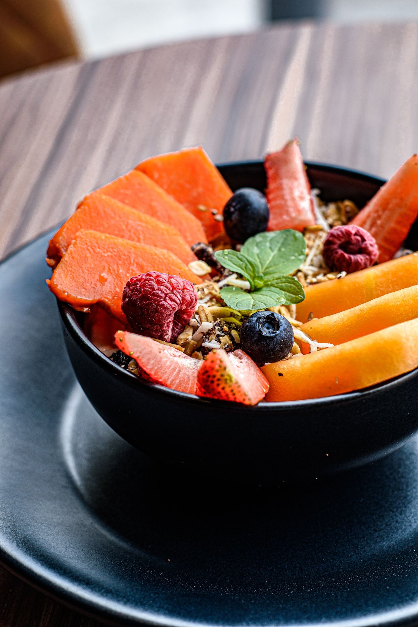 Frutero: papaya, mango, frutos rojos, granola y una ramita de menta. Cuenco negro sobre plato negro, mesa de madera.
