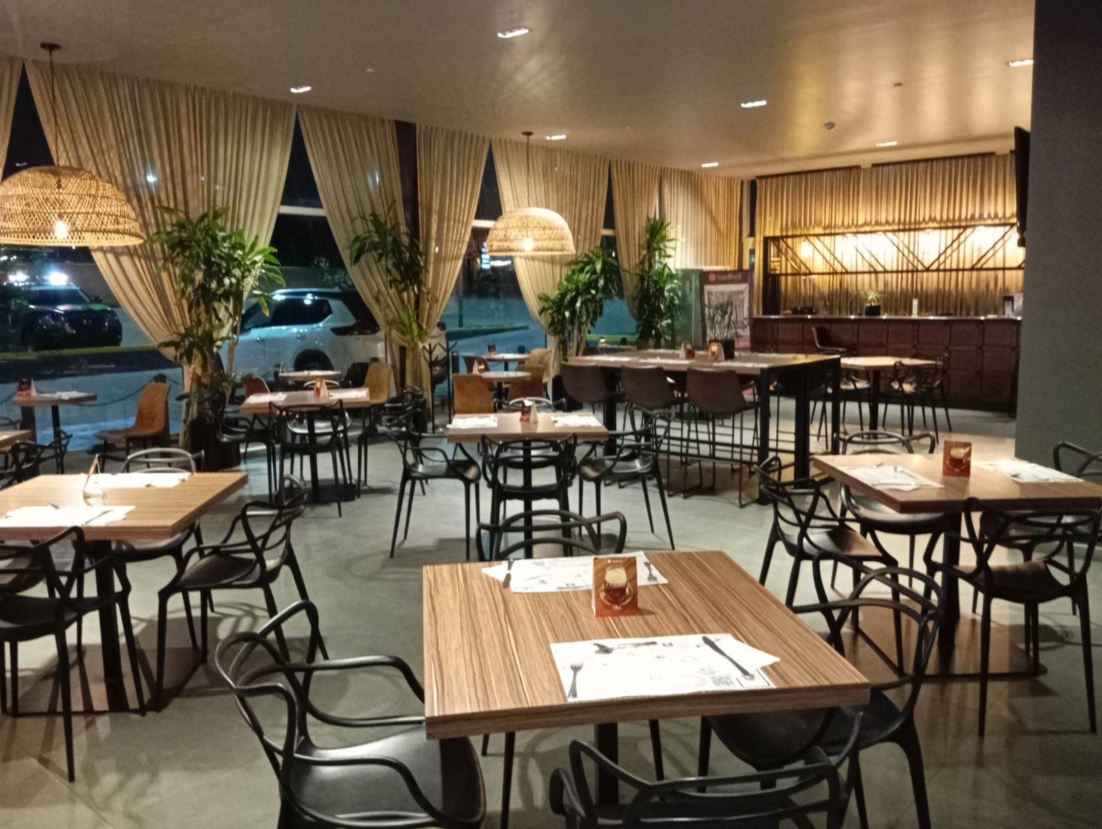 Interior del restaurante con mesas y sillas. Grandes ventanales con cortinas y lámparas colgantes. Zona de bar al fondo.