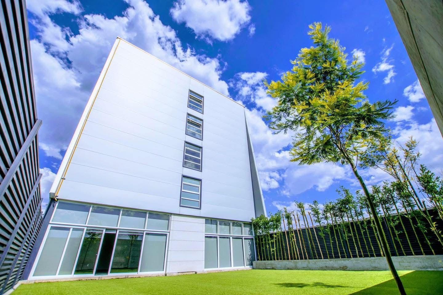 Edificio blanco moderno con fachada de cristal, rodeado de césped y árboles bajo un cielo azul.