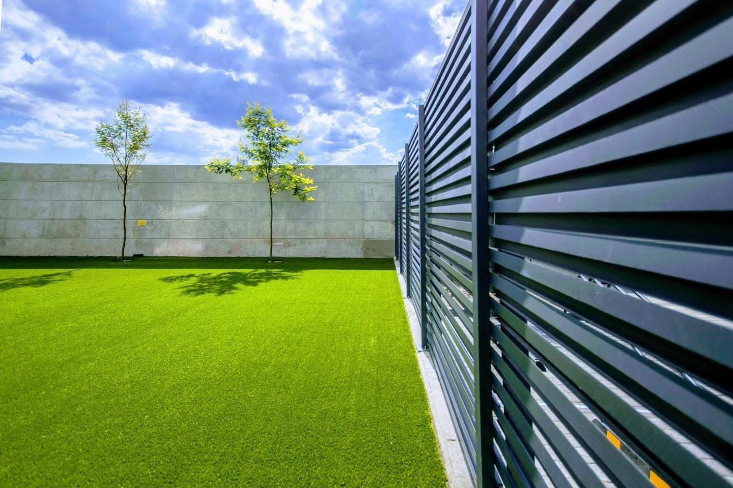 Césped verde y árboles en un patio junto a una valla de listones negros con un cielo azul nublado de fondo.