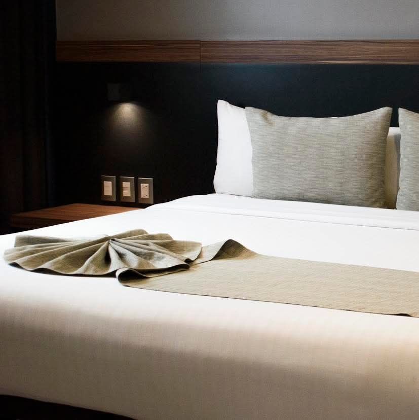 Cama en una habitación de hotel con sábanas blancas, almohadas grises y una toalla doblada.