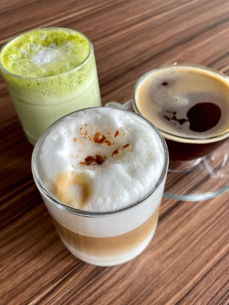 Tres bebidas de café en una mesa de madera: café con leche, café con leche matcha y café negro.