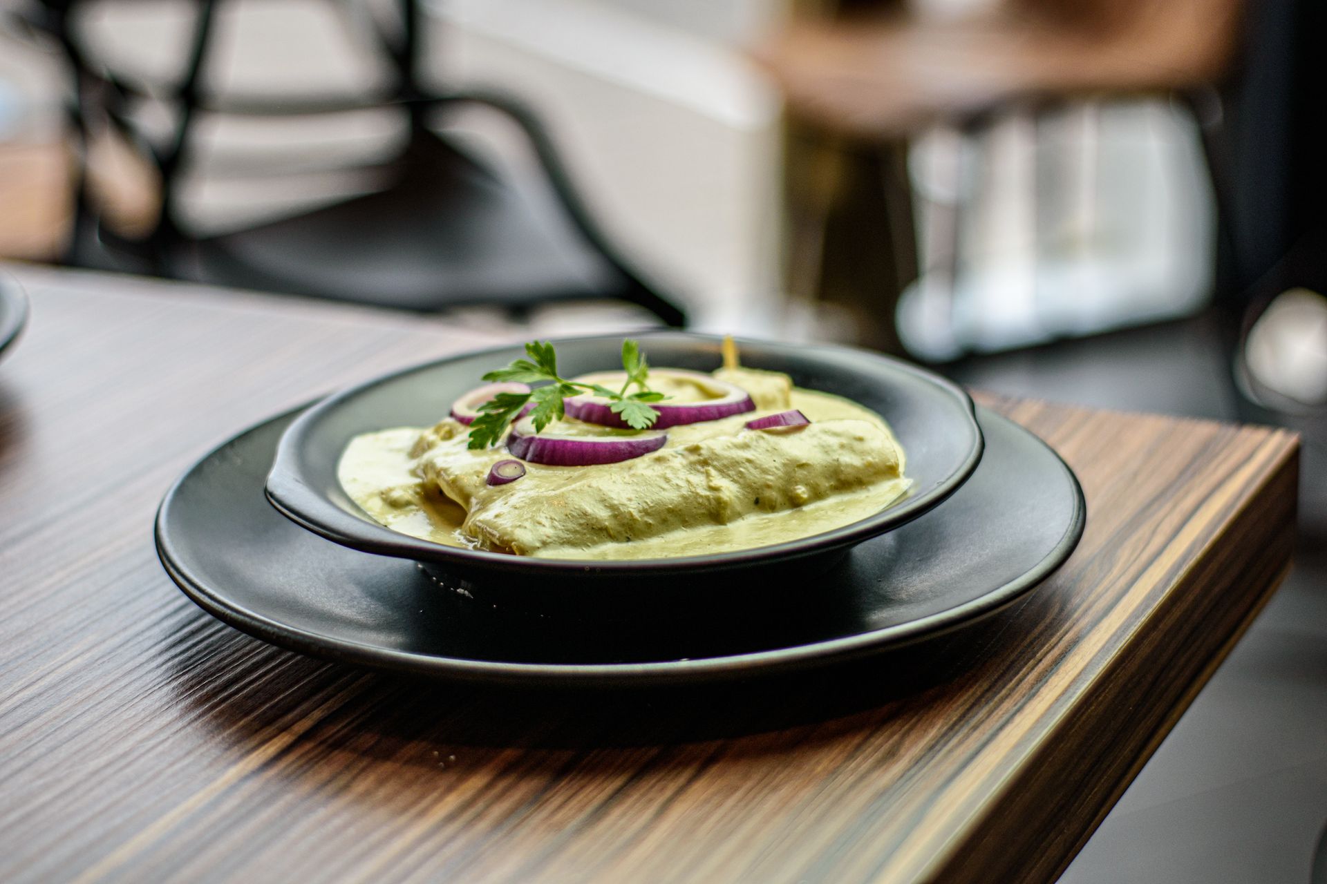 Enchiladas en salsa verde, cubiertas con cebolla roja y perejil, en un tazón negro sobre una mesa.