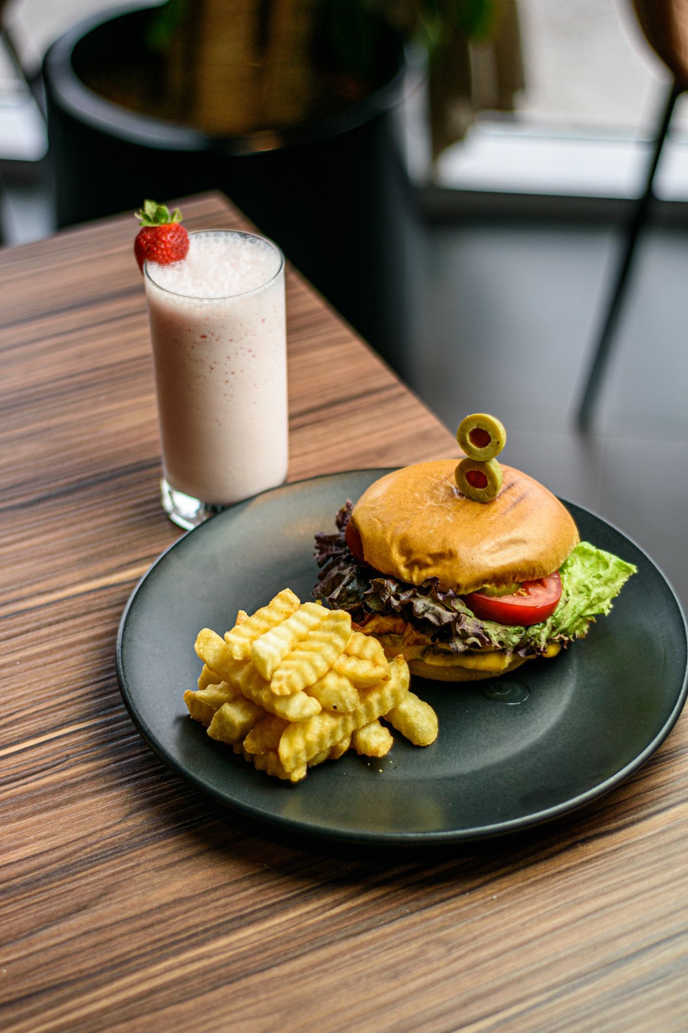 Hamburguesa, patatas fritas y batido de fresa en un plato negro, sobre una mesa de madera.