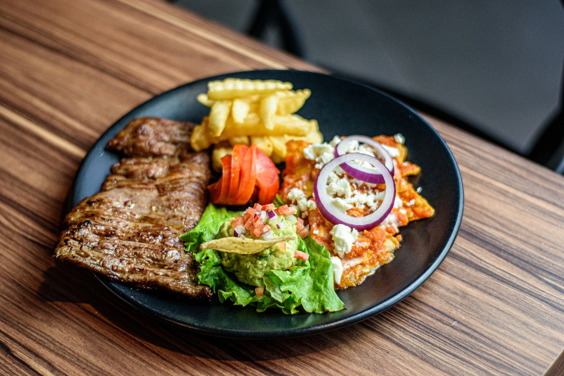 Plato de bistec a la parrilla, chilaquiles con aderezos, papas fritas, guacamole y tomate sobre una mesa de madera.