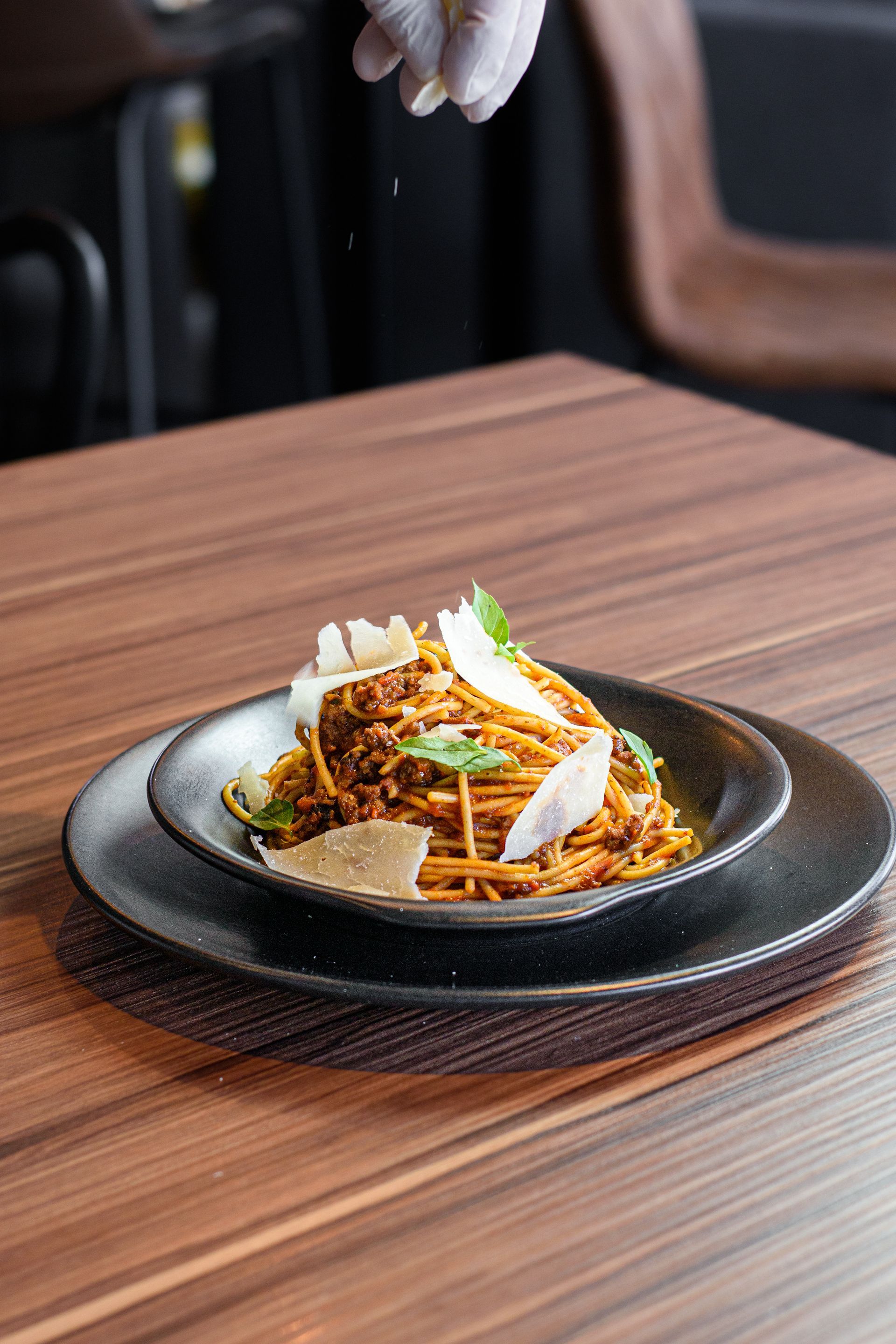 Espaguetis con salsa y parmesano, condimentados con una mano enguantada, servidos en un plato negro sobre una mesa de madera.