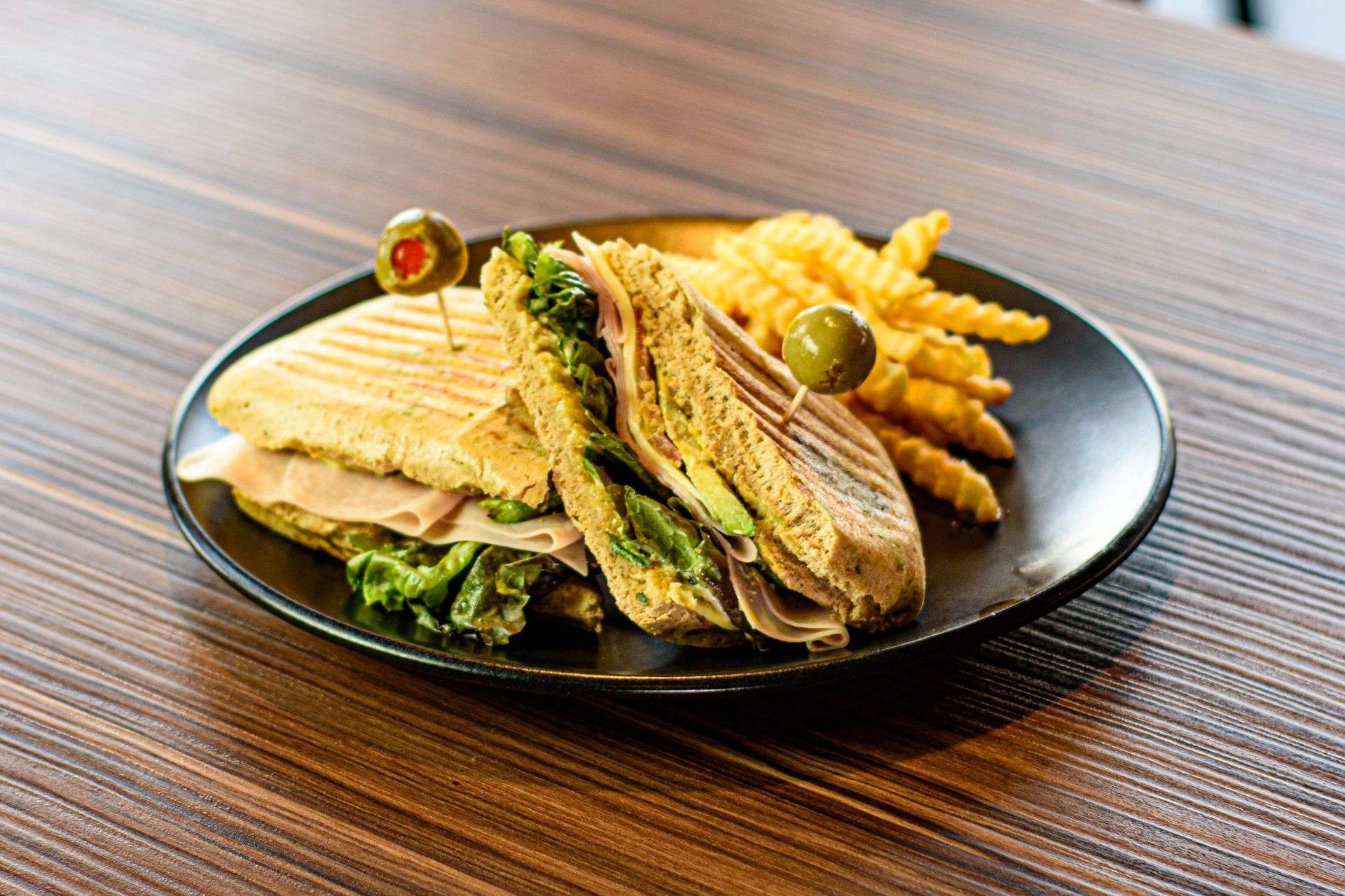 Sándwich panini con lechuga, jamón, aguacate y papas fritas onduladas en un plato negro, adornado con aceitunas.