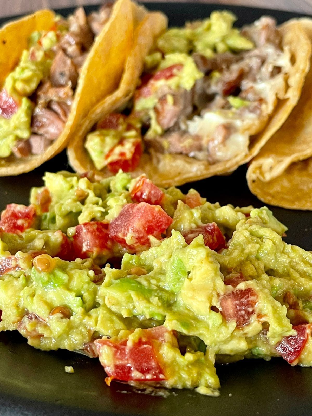 Tacos con guacamole en un plato negro. Los tacos están rellenos de carne y cubiertos con aguacate y tomate.