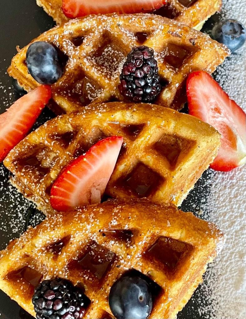 Waffles cubiertos con fresas, arándanos, moras y azúcar en polvo en un plato negro.