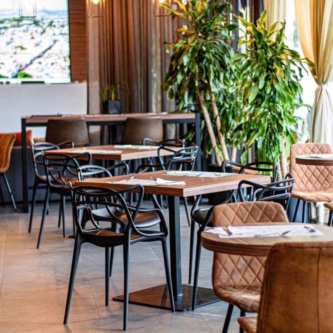 Interior del restaurante: mesas con sillas, plantas altas y verdes, paleta de colores neutros.