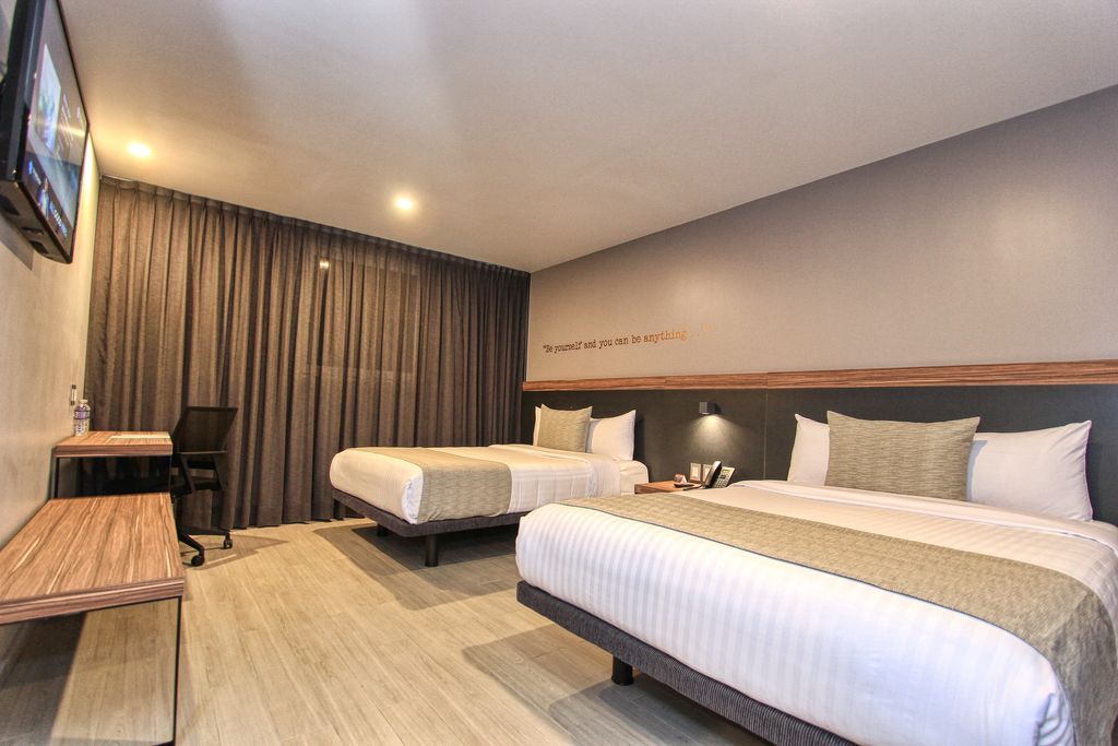 Habitación de hotel con dos camas, una doble y una individual, detalles de madera, cortinas grises y escritorio.