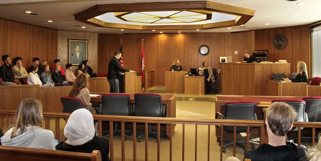 a group of people are sitting in a courtroom watching a man give a speech .